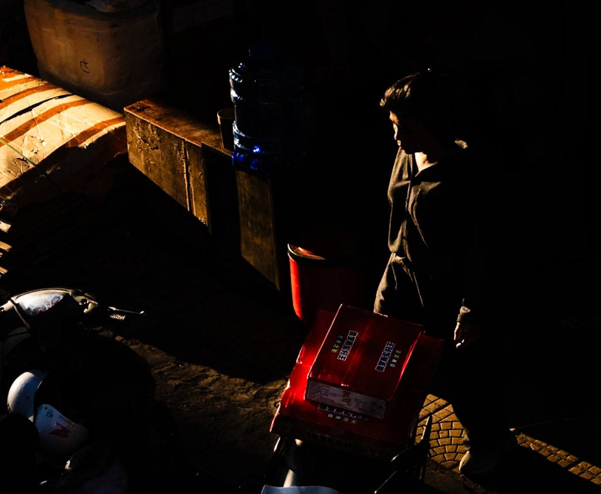 A figure wheels a red crate of goods through a narrow shaft of light in an otherwise dark yard — Jim Kerns
