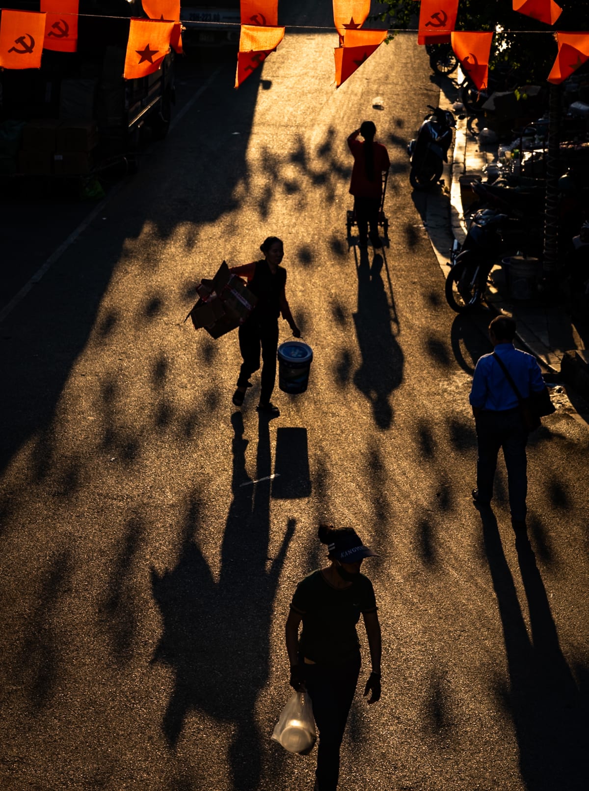 Seen from above, silhouetted pedestrians carrying boxes, a paint bucket and a plastic bag cross a low-sun street strung with red flags and long tree shadows — Jim Kerns