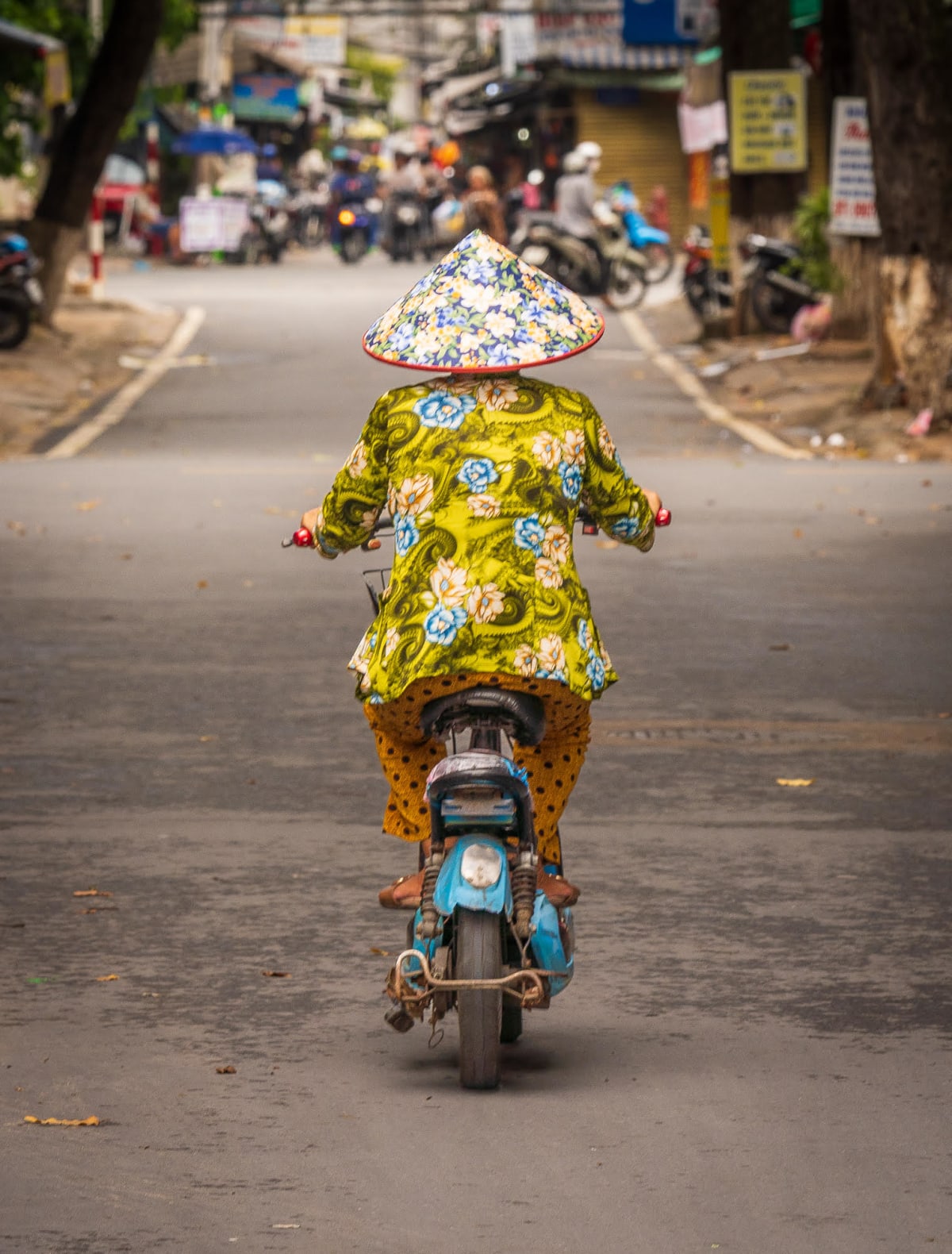 A woman in a floral conical hat and a matching yellow-green floral blouse rides a battered blue motorbike away down the middle of a quiet street, the traffic and shopfronts ahead of her falling out of focus — Jim Kerns
