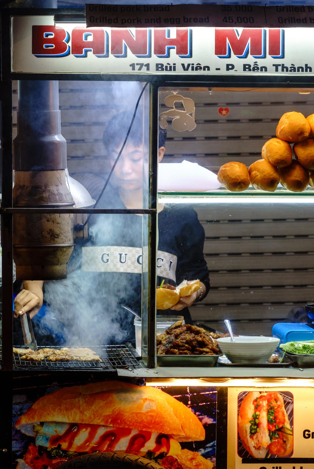 Behind the lit glass of a bánh mì cart on Bùi Viện, one cook turns pork on the grill in a cloud of smoke while another fills a split baguette, a pile of rolls glowing on the shelf above — Jim Kerns