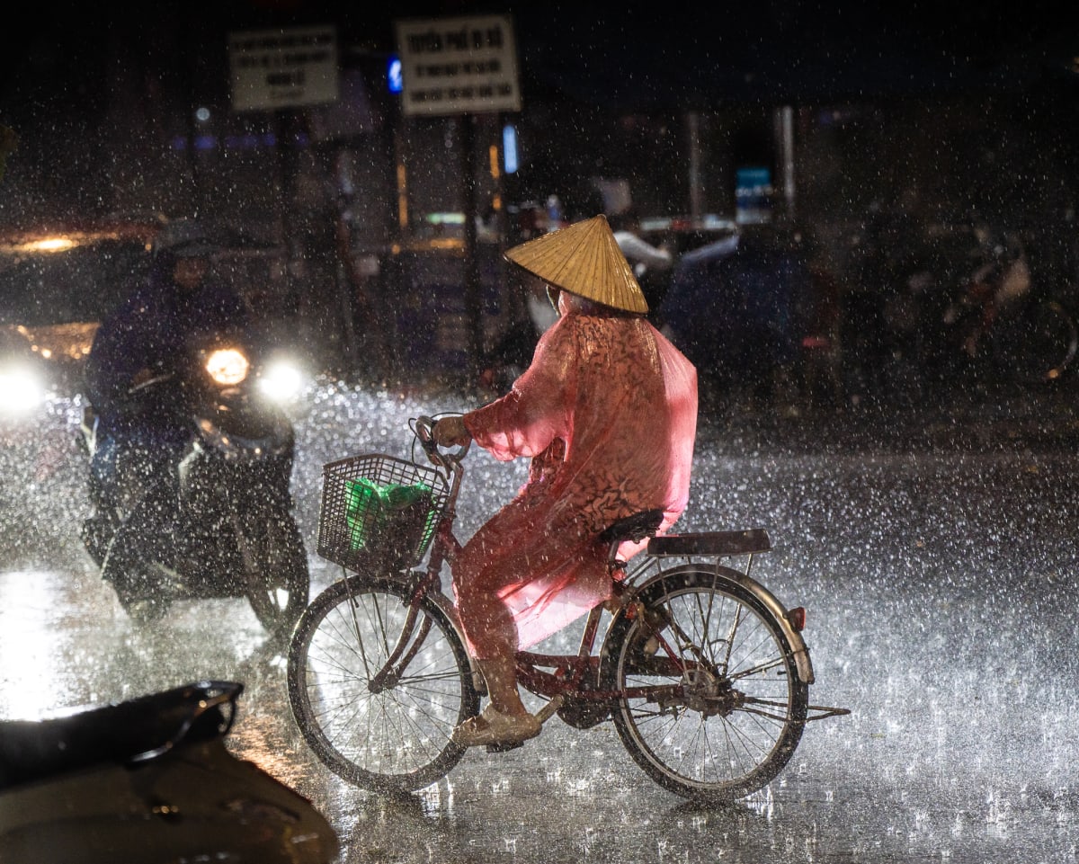 A woman in a pink plastic poncho and a gold conical hat pedals a bicycle across a junction in heavy night rain, headlights behind her catching every falling drop — Jim Kerns