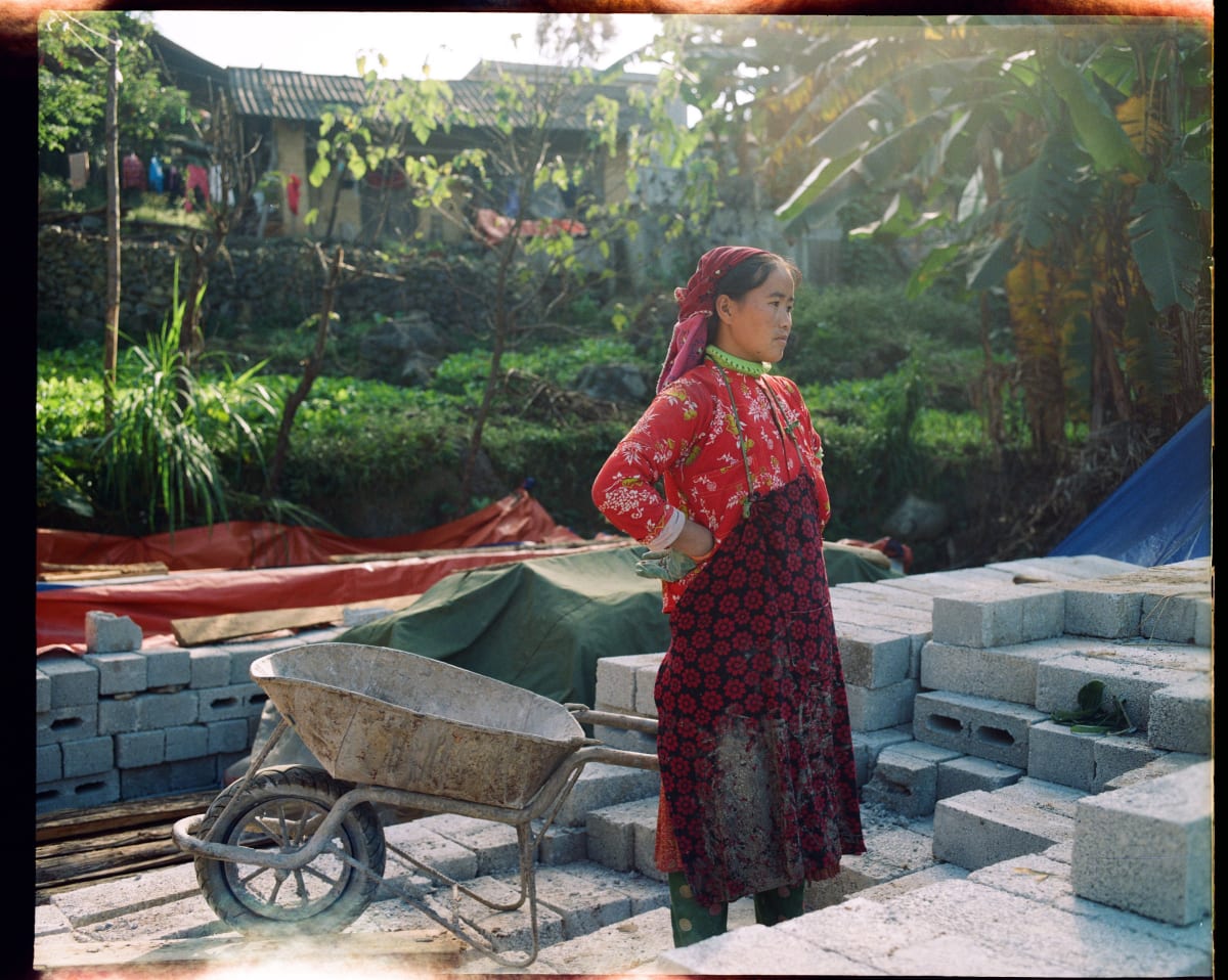 A young woman in a red embroidered jacket stands with her hands on her hips beside a wheelbarrow and stacks of concrete blocks, a house going up behind her — Dennis and Patrick Weinert