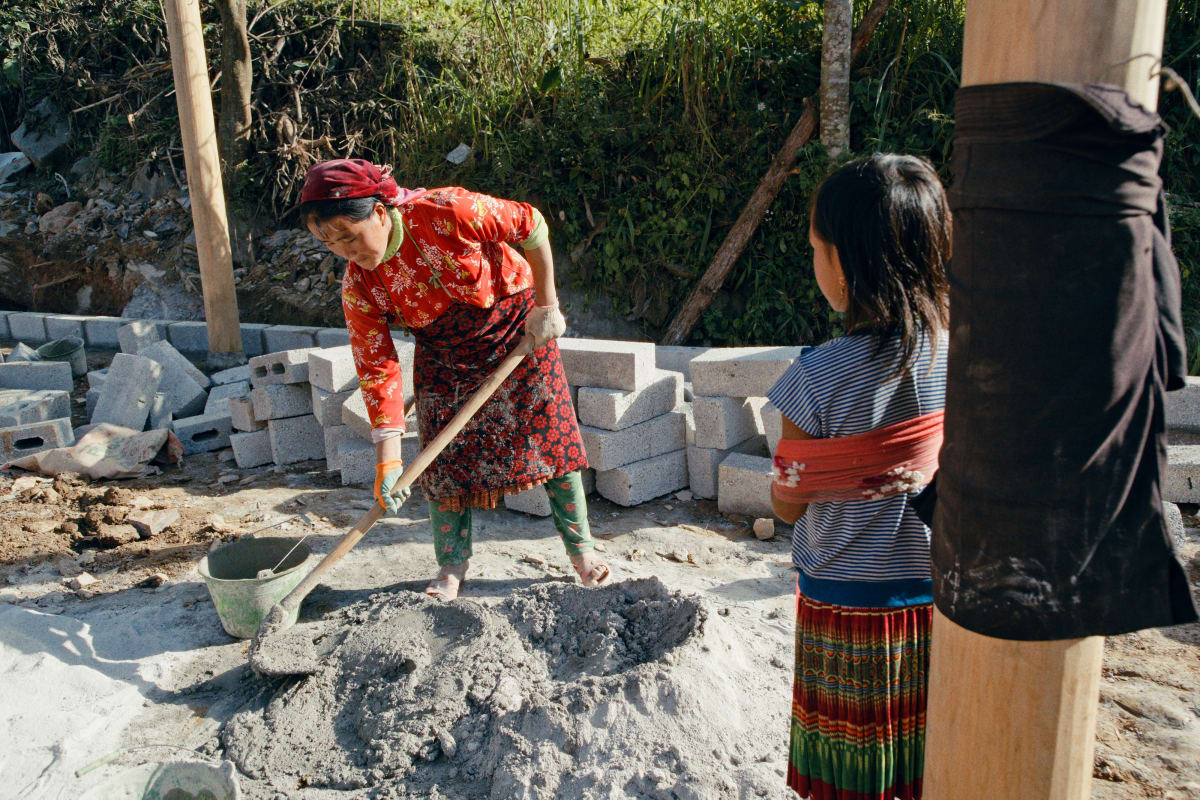 A woman in a red jacket turns mortar with a hoe while a girl in a pleated skirt, her arm in a sling, watches from beside a timber post — Dennis and Patrick Weinert
