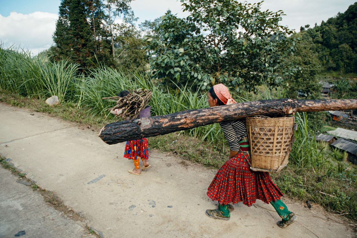 A woman carries a whole timber beam on her shoulder along a concrete path, a woven basket on her back and a small child walking ahead with a bundle of sticks — Dennis and Patrick Weinert