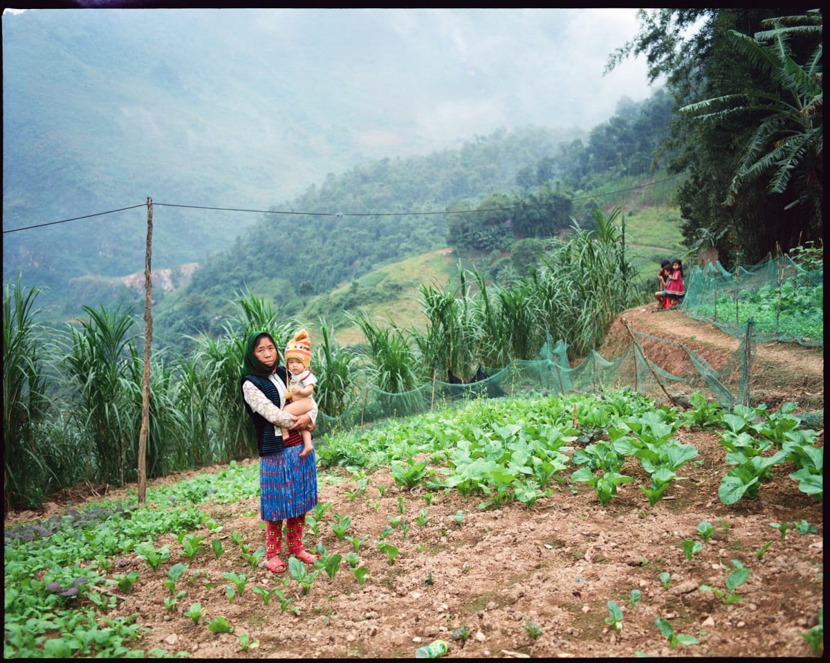 A young woman holds a baby in a knitted bonnet at the edge of a vegetable plot, terraced green mountains rising into mist behind her — Dennis and Patrick Weinert