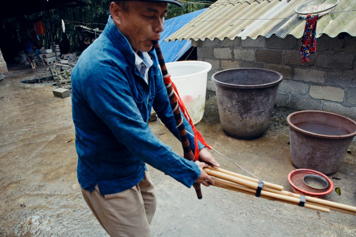 A man in an indigo jacket crosses a concrete yard playing the khèn, water basins and a corrugated roof behind him — Dennis and Patrick Weinert