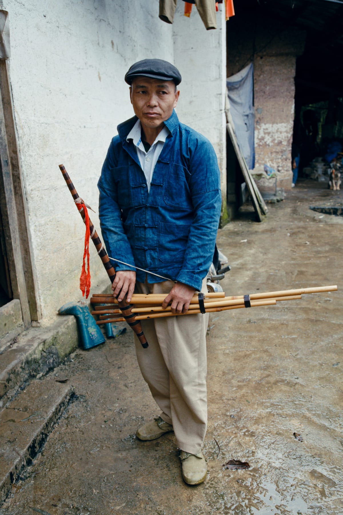 A man in an indigo jacket and flat cap stands holding his khèn across his body, looking into the camera — Dennis and Patrick Weinert