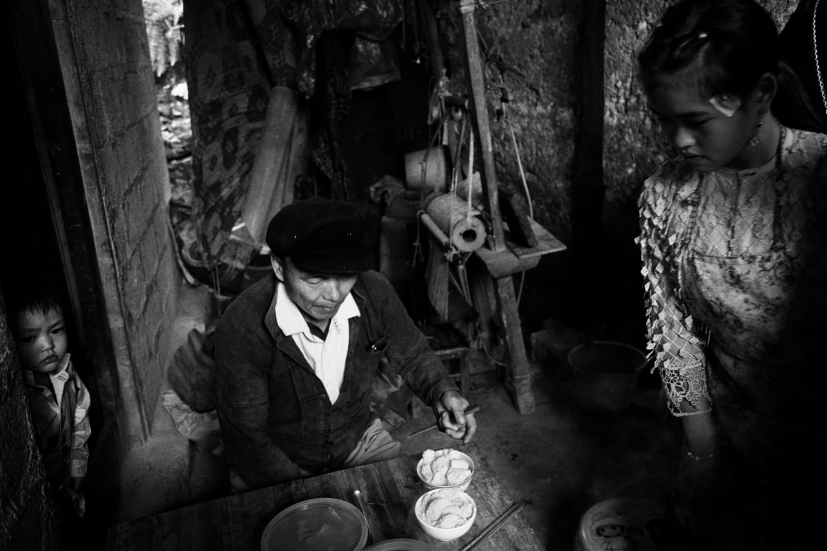 A man in a flat cap sits at a low table set with bowls in a dark room, a young woman standing at his shoulder and a small child watching from the doorway — Dennis and Patrick Weinert
