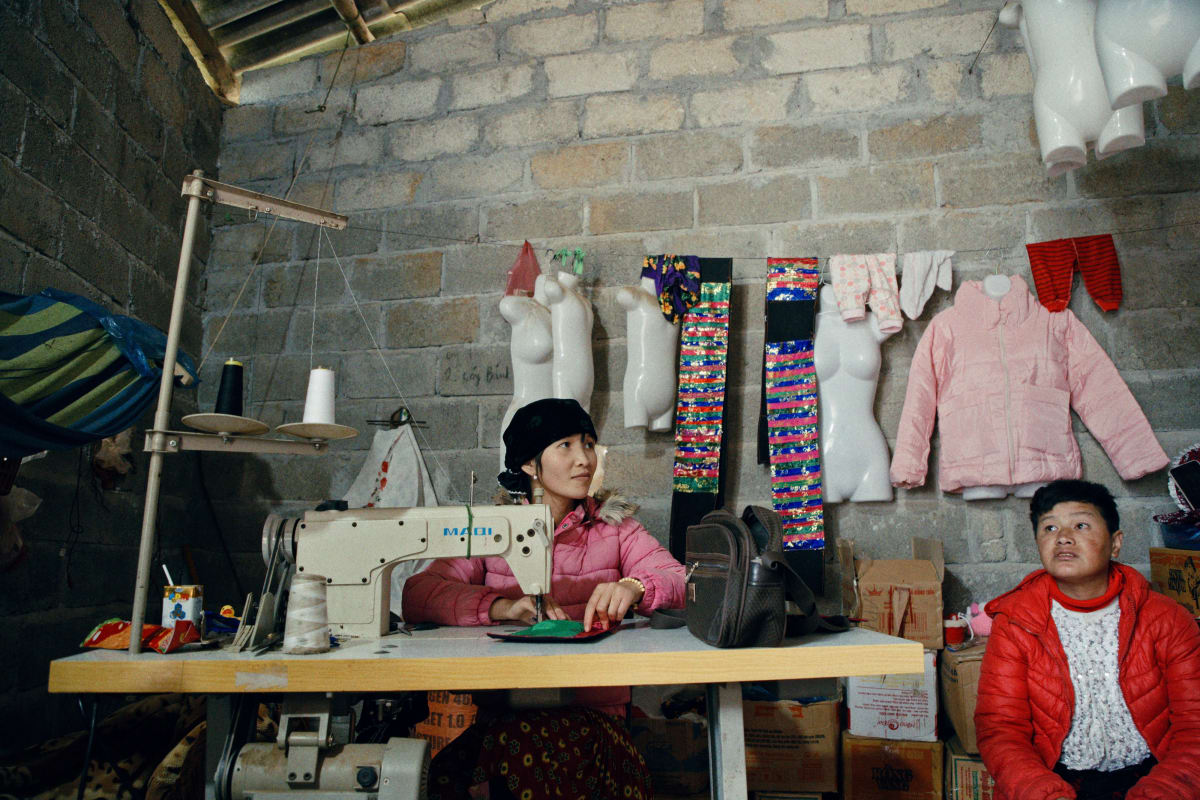 A woman works an industrial sewing machine against a breeze-block wall hung with mannequin torsos, bright leg-wraps and a child's pink coat, a boy in red sitting beside her — Dennis and Patrick Weinert
