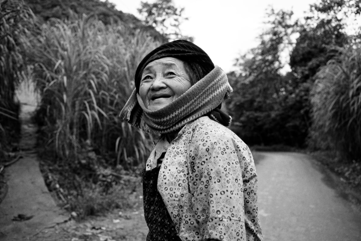 An older woman in a headwrap and patterned jacket turns on a road to smile at the camera, tall grass behind her — Dennis and Patrick Weinert