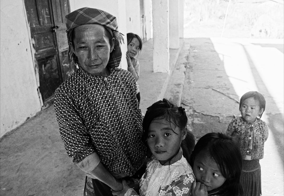 A woman in a checked headwrap stands on a concrete veranda with four children gathered around her, all looking at the camera — Dennis and Patrick Weinert