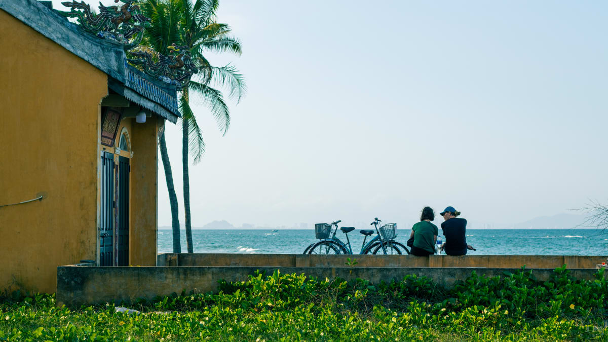 Two friends sit with bicycles by a Vietnamese coastal temple under palm trees gazing at the sea