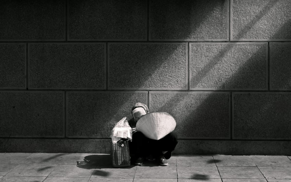 Vietnamese woman with conical hat crouches beside a wall in a shaft of light, quiet and solitary