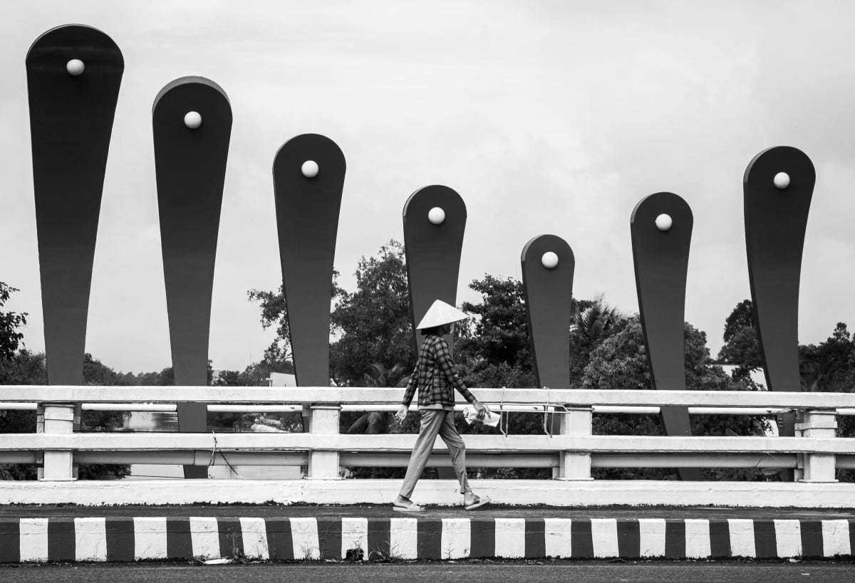 Woman in conical nón lá hat walks across a modern Vietnamese bridge with tall decorative lampposts