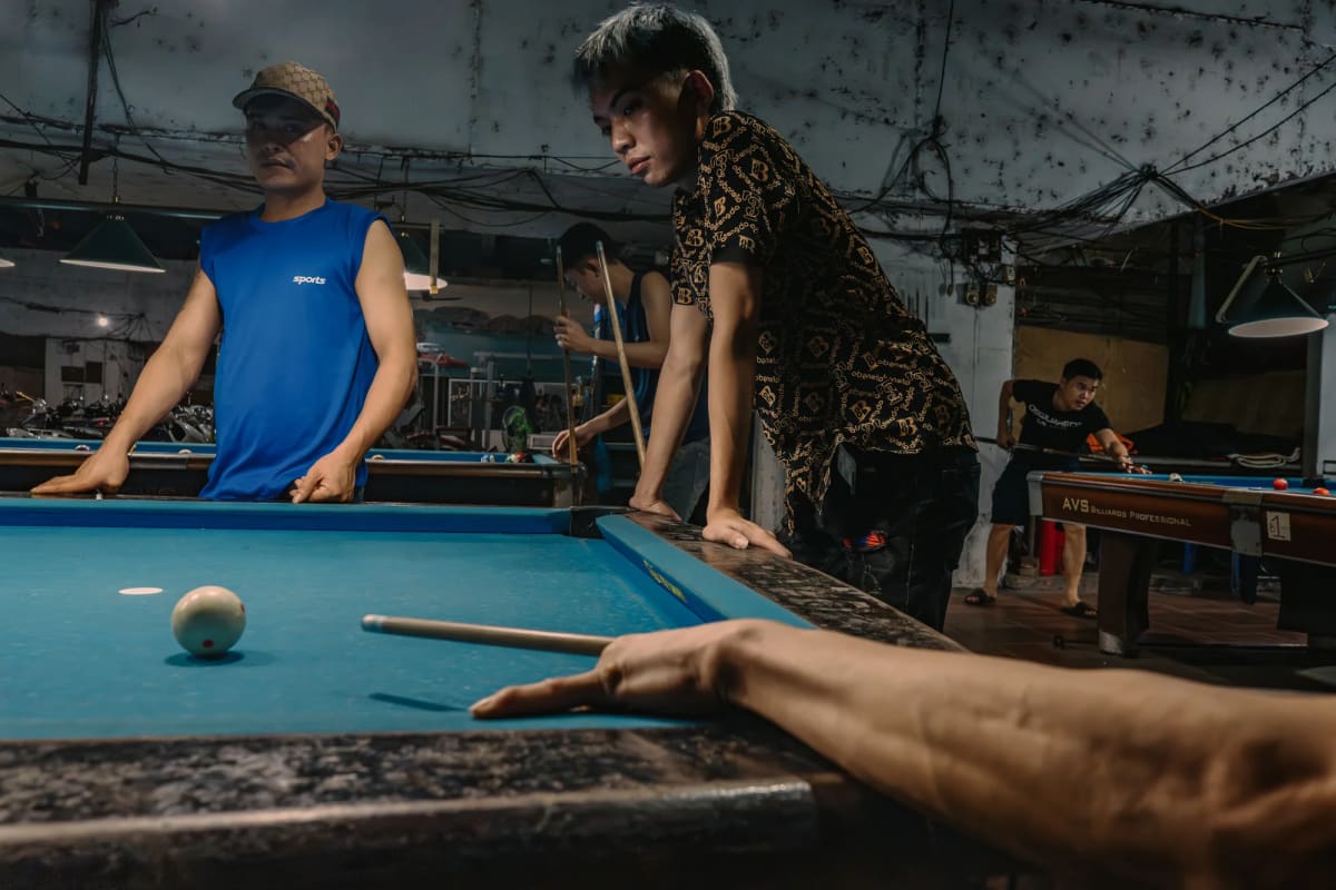 Three young men at a blue-felt pool table in a dim Hanoi billiards hall; the central player in a black-and-gold patterned shirt leans into his shot while a man in a blue sleeveless vest watches from the left, and a foreground hand and cue enter from the lower right — Antonio di Vico