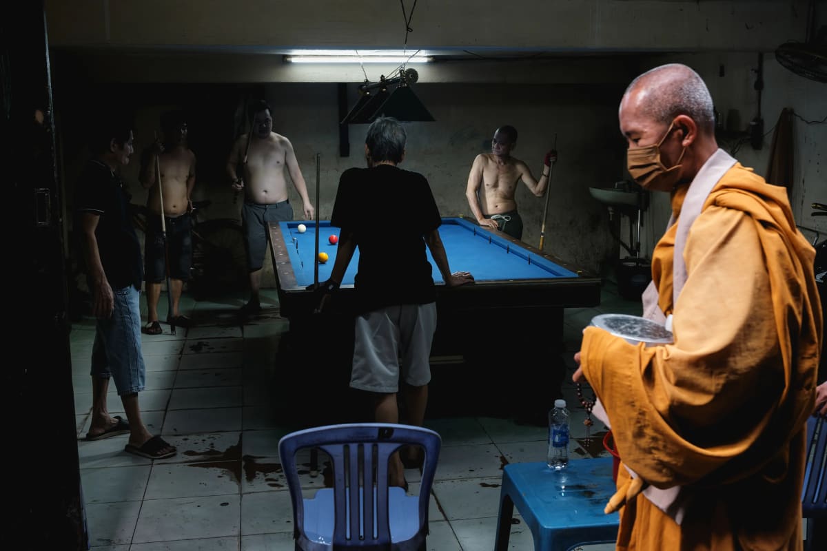 A saffron-robed, masked monk holding an alms bowl and prayer beads passes through a dim backstreet pool hall where shirtless men lean on cues around a blue-felt table — Adrien Jean.