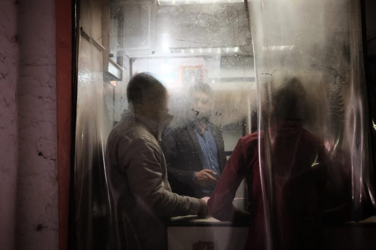 Three men stand at a night food stall, their figures softened by a scratched sheet of hanging plastic between them and the camera — Luke Neville