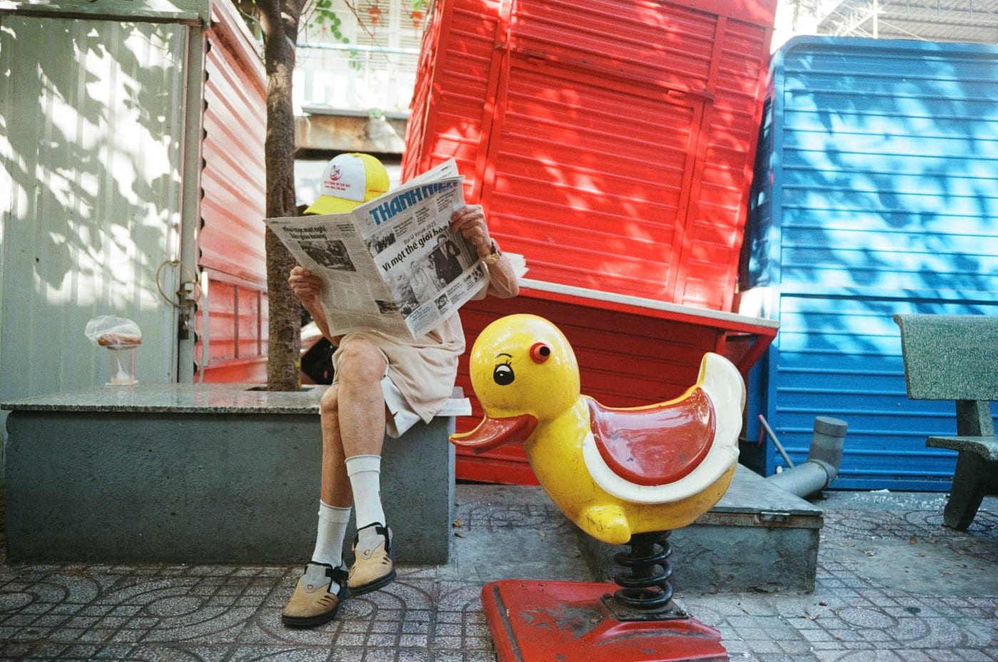 Man reads newspaper beside a yellow duck ride — Le Hoang Tuan