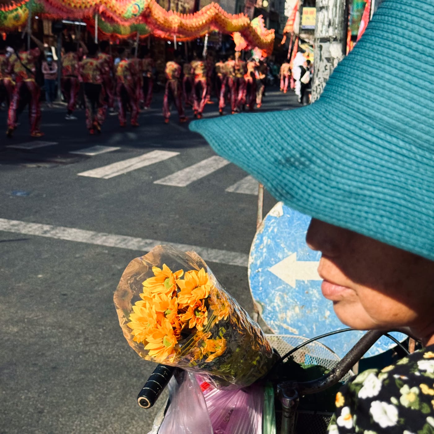 Flower seller in a teal hat watches a dragon dance pass — Le Hoang Tuan