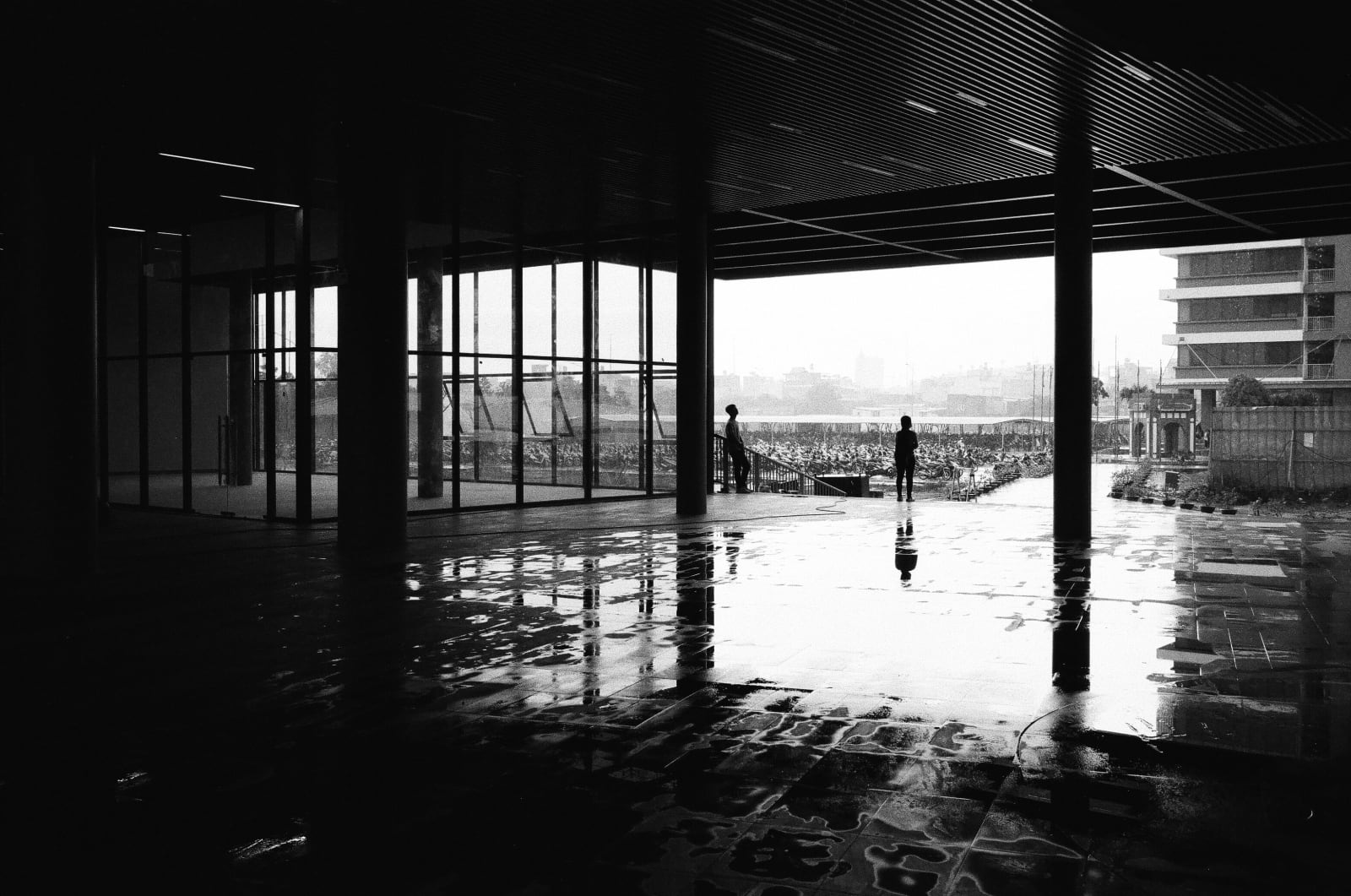 Two figures silhouetted at the edge of a covered concrete walkway, looking out over a rain-wet plaza toward rows of parked motorbikes and a hazy city skyline