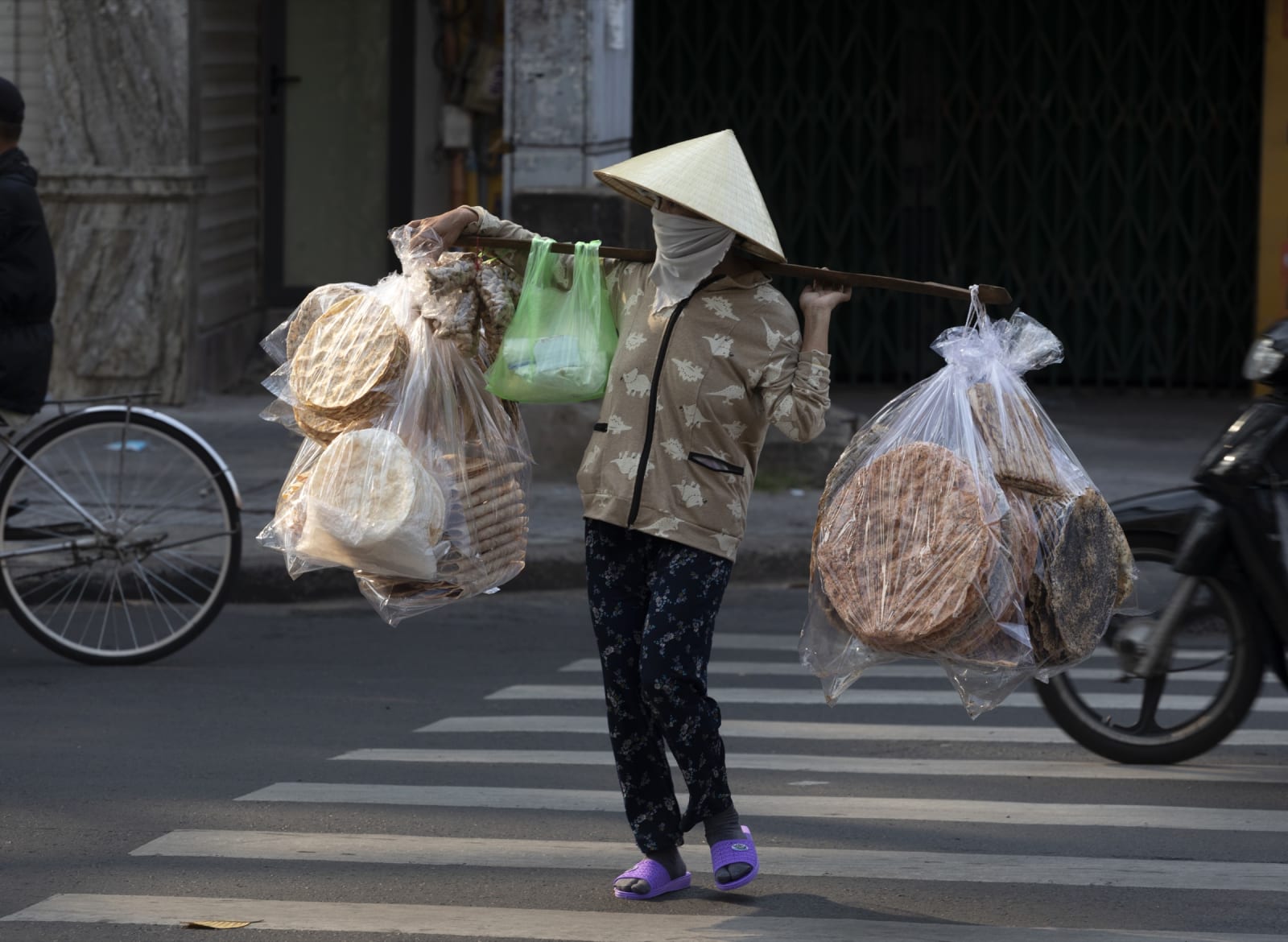A rice paper vendor in a conical hat carries stacked bags of bánh tráng across a Saigon street