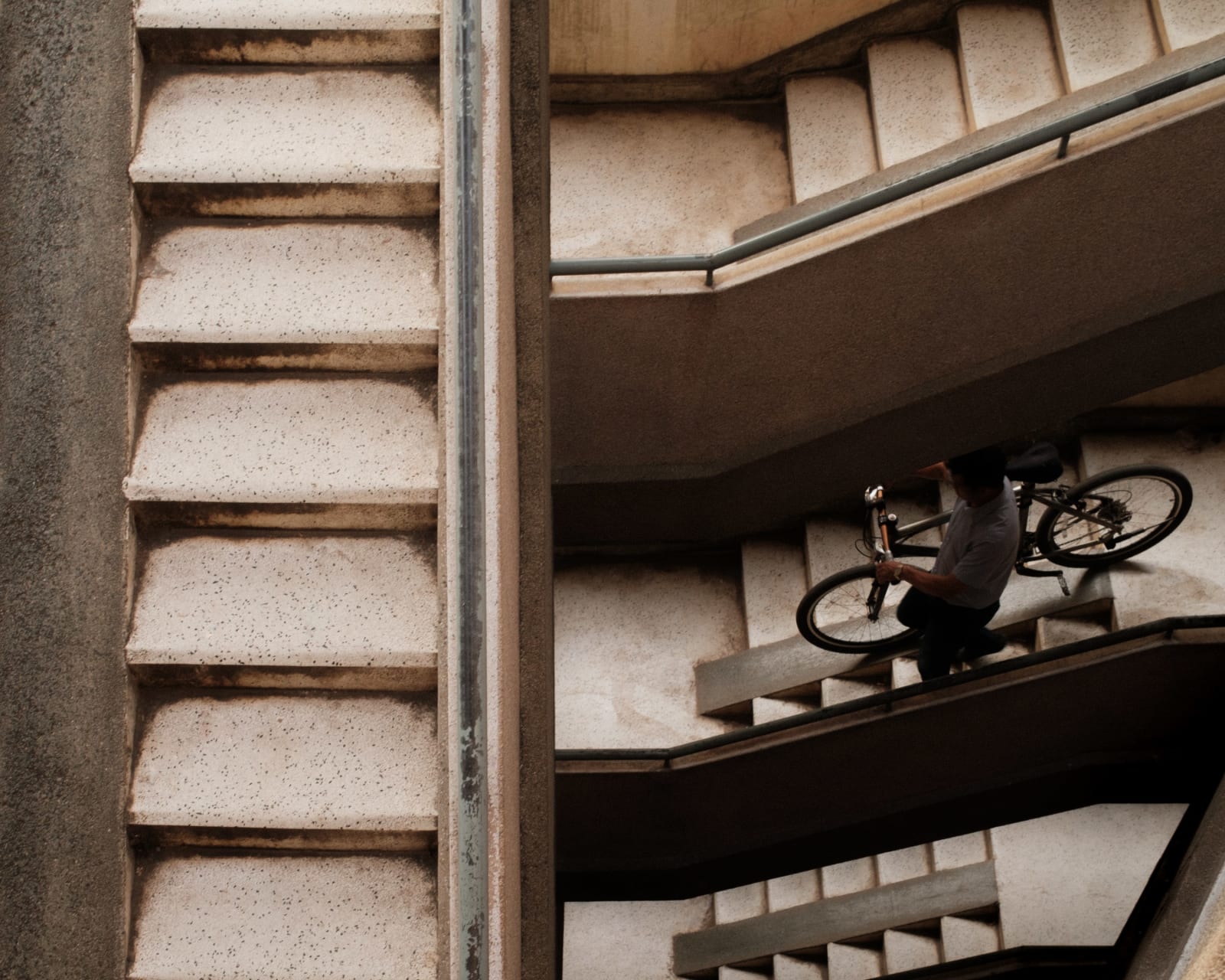 Seen from above, a man carries a bicycle down a concrete apartment stairwell