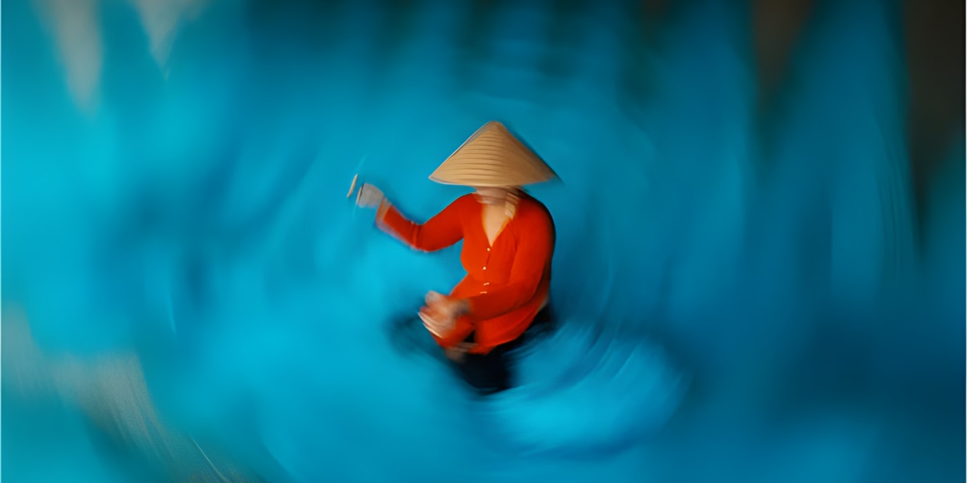 A woman in a red áo bà ba and conical nón lá hat sits in a round basket boat at the center of the frame, surrounded by vivid turquoise water rendered into a radial vortex of blur, her hand raised slightly — Jimmy Ky