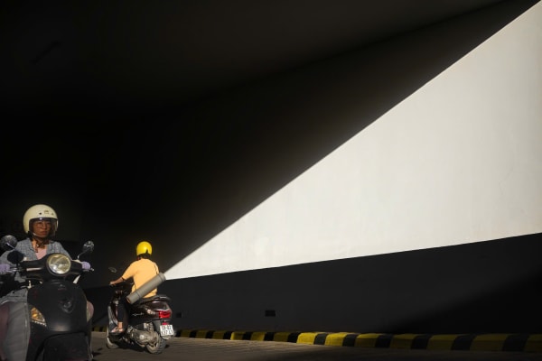 Motorbikes pass under a tunnel as a sharp diagonal of sunlight cuts across the wall — MatDat.Ng