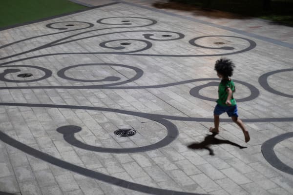 A barefoot child in a green shirt runs at night across a plaza inlaid with dark swirling scrollwork, casting a long shadow — Paul Evan Green