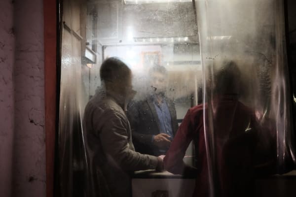 Three men stand at a night food stall, their figures softened by a scratched sheet of hanging plastic between them and the camera — Luke Neville