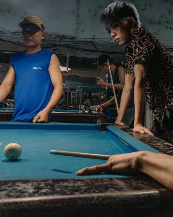 Three young men at a blue-felt pool table in a dim Hanoi billiards hall; the central player in a black-and-gold patterned shirt leans into his shot while a man in a blue sleeveless vest watches from the left, and a foreground hand and cue enter from the lower right — Antonio di Vico