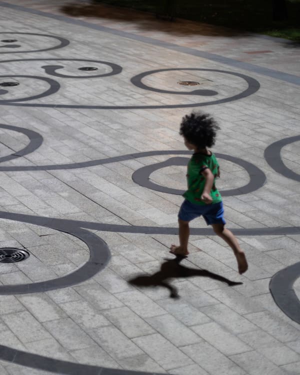 A barefoot child in a green shirt runs at night across a plaza inlaid with dark swirling scrollwork, casting a long shadow — Paul Evan Green
