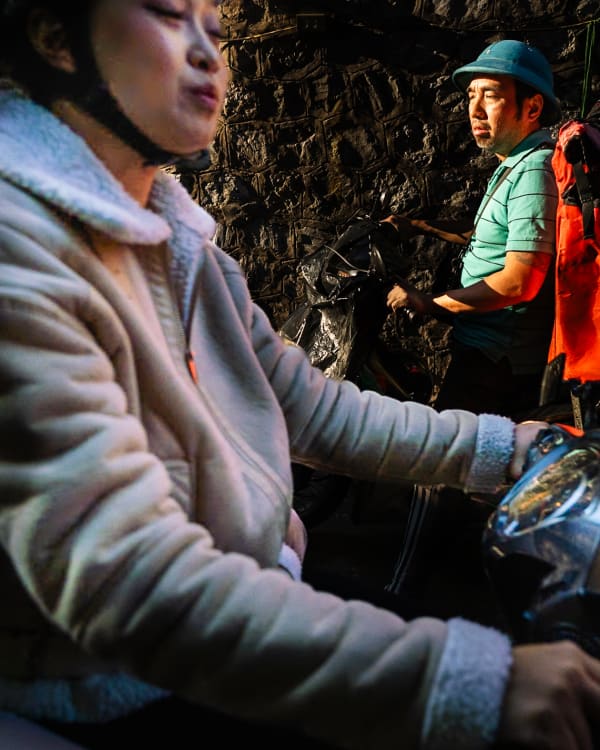 A woman on a scooter and a delivery rider with an orange cargo box pass one another in a dark stone alley — Jim Kerns