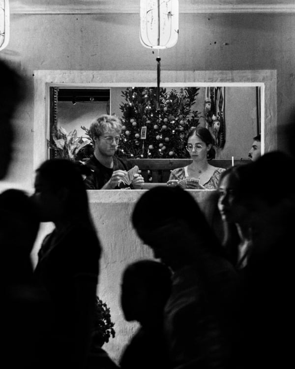 Couple playing cards framed through a window among silhouetted crowds at a Hoi An cafe in black and white