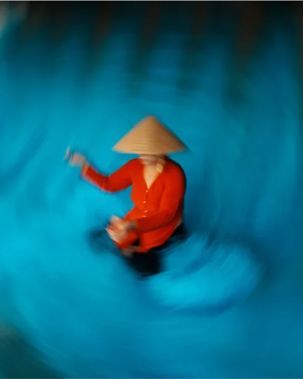 A woman in a red áo bà ba and conical nón lá hat sits in a round basket boat at the center of the frame, surrounded by vivid turquoise water rendered into a radial vortex of blur, her hand raised slightly — Jimmy Ky