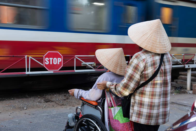 Two women in conical hats wait at a railway crossing behind a red stop sign as a blue-and-red train blurs past, one standing with her hand on her companion’s wheelchair — Paul Evan Green