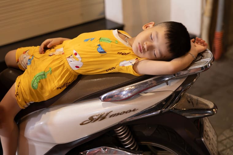 A young boy in a yellow shirt sleeps stretched out on the seat of a parked scooter, arms folded behind his head — Paul Evan Green
