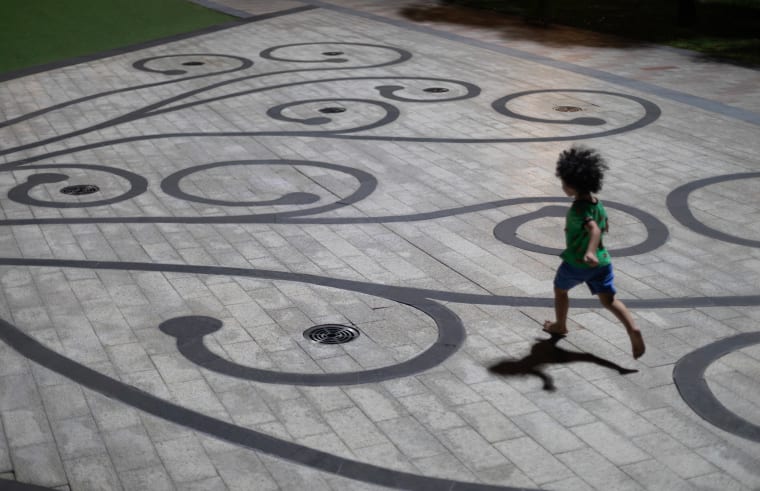 A barefoot child in a green shirt runs at night across a plaza inlaid with dark swirling scrollwork, casting a long shadow — Paul Evan Green