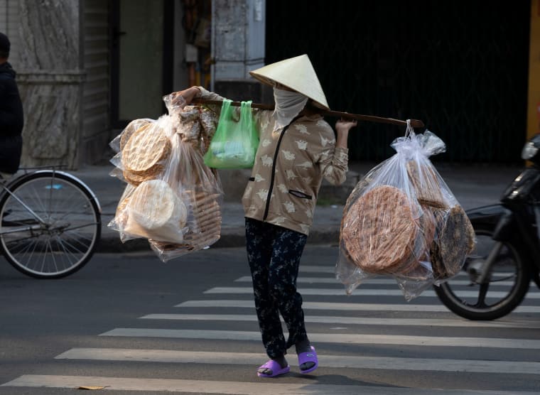 A street vendor in a green conical hat crosses a zebra crossing under a shoulder pole strung with bags of rice crackers — Paul Evan Green