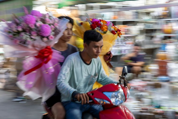 Two men on a motorbike carry large wrapped flower bouquets through a market street, the background streaked with motion — Paul Evan Green