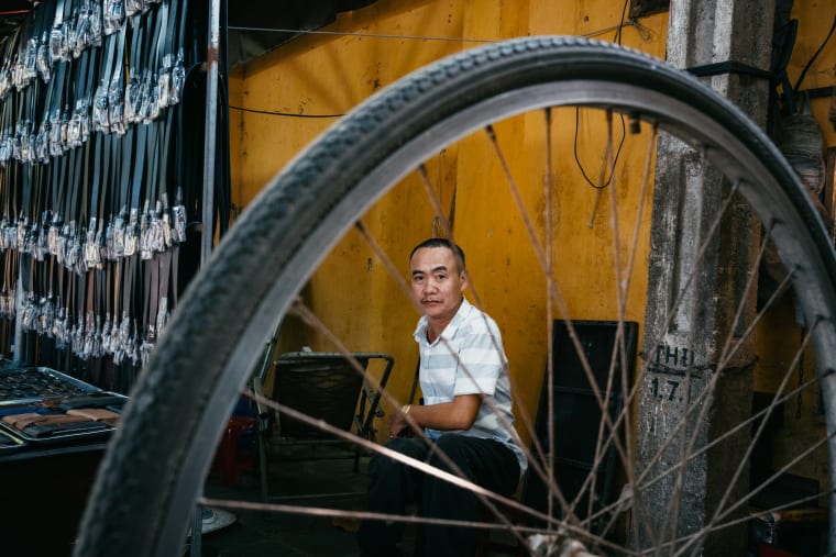 Framed through a bicycle wheel's spokes, a man in a striped shirt sits watching against a weathered yellow wall hung with goods — Antonio di Vico