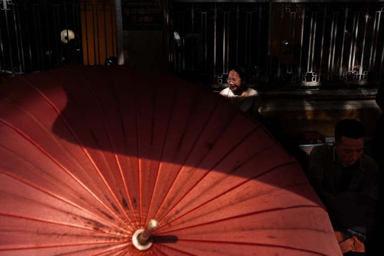 Sunlight rakes across a large red silk parasol as a bespectacled woman smiles and a man works in the shadowed background — Antonio di Vico