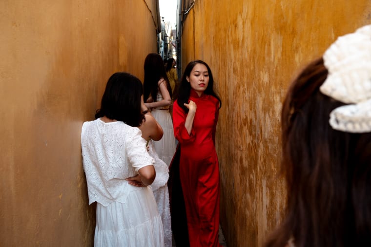Women in white and a vivid red ao dai pose along a narrow, sunlit passage between weathered ochre walls — Antonio di Vico
