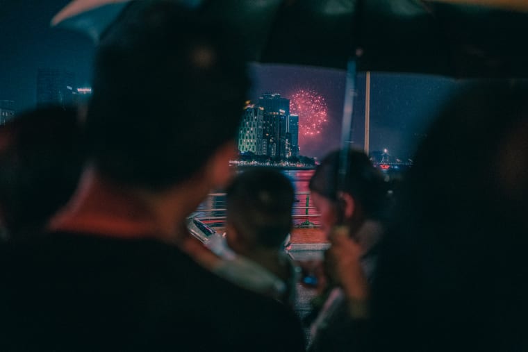Silhouetted spectators watch pink fireworks burst over a riverfront skyline and lit high-rise on a hazy night — Antonio di Vico