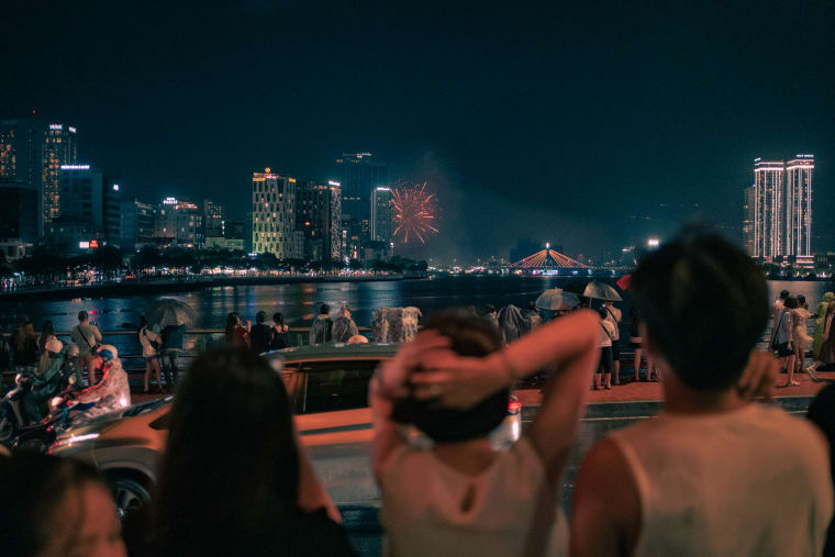 A nighttime crowd with umbrellas lines a rain-slicked riverbank watching orange fireworks explode above the illuminated city skyline and bridge — Antonio di Vico