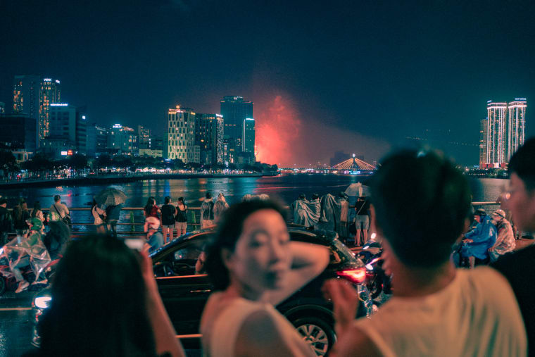 Night crowds in rain ponchos watch red fireworks bloom over a river beside a lit cable-stayed bridge and city towers — Antonio di Vico