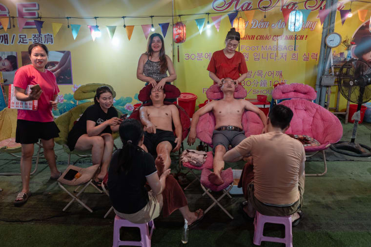 Two shirtless men get head and foot massages in reclining chairs at a bright yellow-lit night-market stall strung with pennant flags — Antonio di Vico