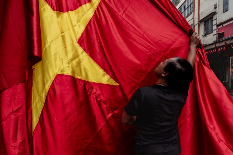 A man in a black T-shirt reaches up to adjust an enormous red Vietnamese flag with a yellow star hanging over an Old Quarter street — Antonio di Vico