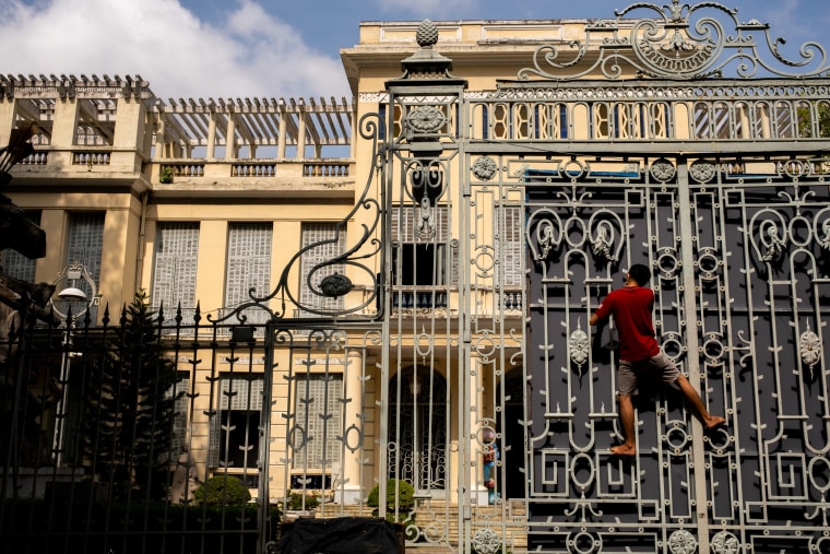 A barefoot man in red climbs the ornate ironwork of a colonial palace gate — Luke Neville