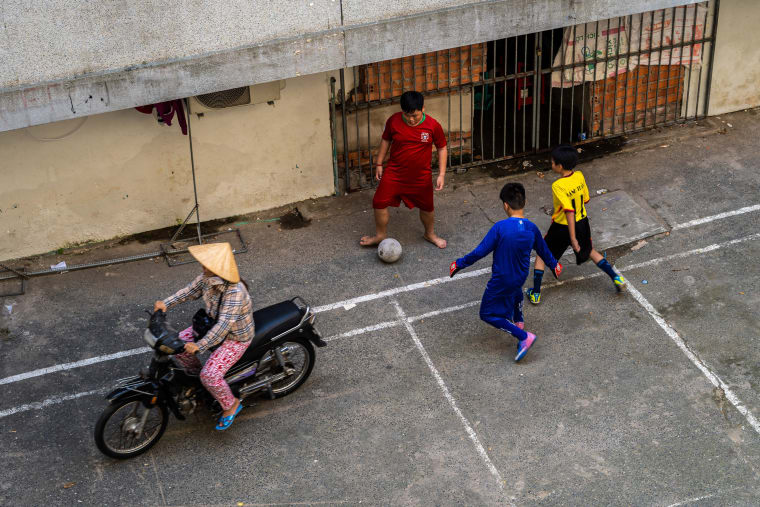 Seen from above, boys play football on a painted court as a woman in a conical hat rides past on a motorbike — Luke Neville